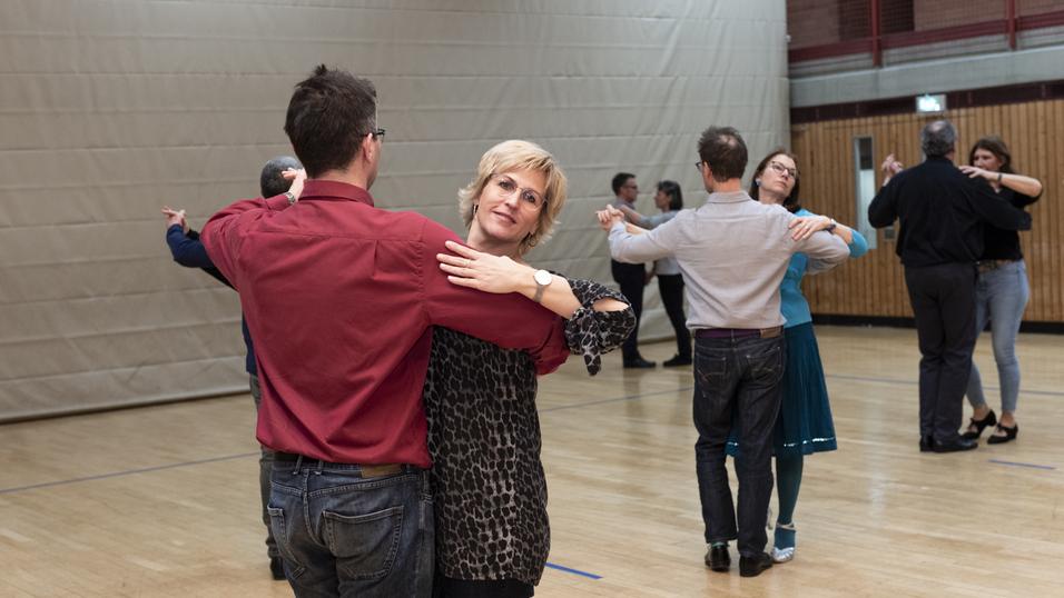 Tanzpaare &uuml;ben im Gymnastikraum, eine Frau in Leo-Bluse tanzt mit einem Mann in rohem Hemd.