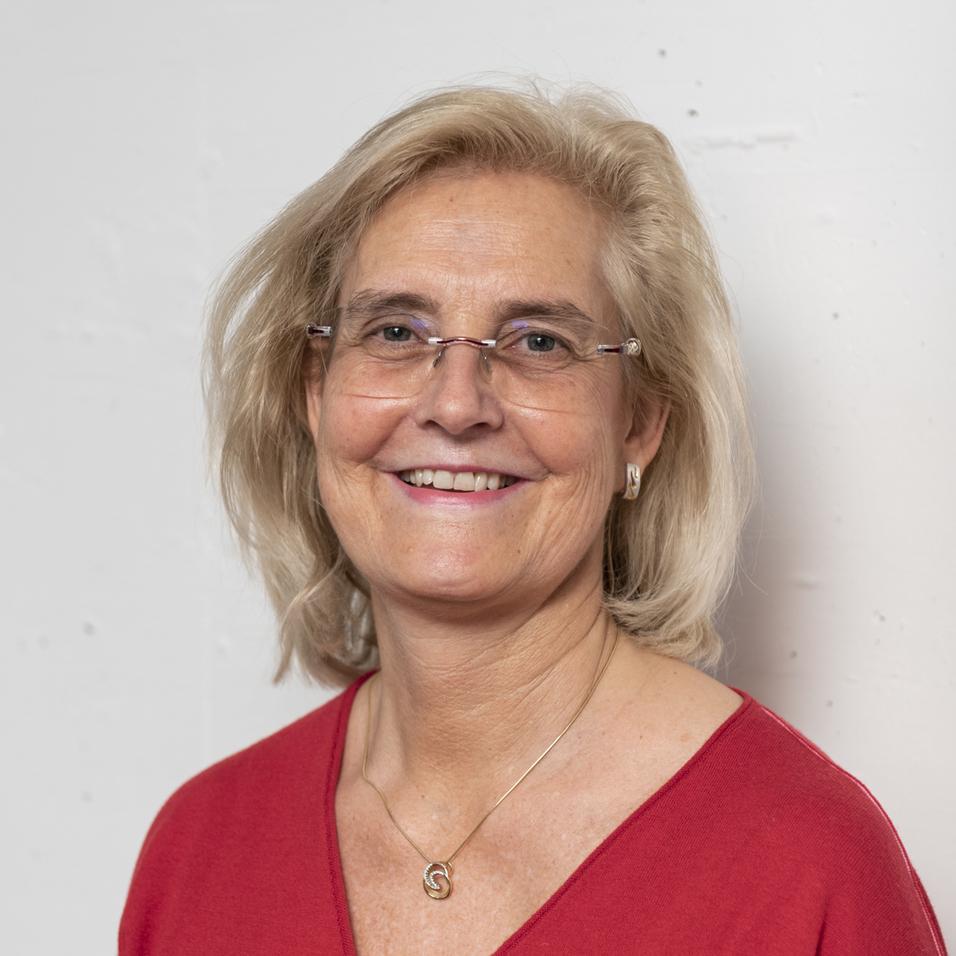 Portrait einer l&auml;chelnden Frau mit blonden, lockigen Haaren, die ein rotes Oberteil tr&auml;gt, vor hellem Hintergrund.