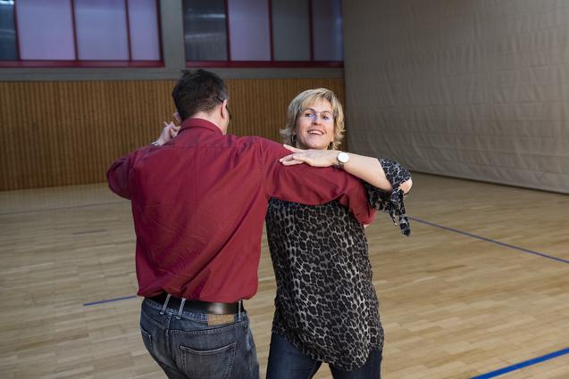 Tanzendes Paar in einem Sportraum, Frau trägt ein geblümtes Oberteil, Mann ein rotes Hemd, beide lächeln.
