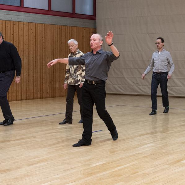 Gruppentanz von sieben M&auml;nnern in einer Sporthalle mit Holzfu&szlig;boden und neutralem Hintergrund.