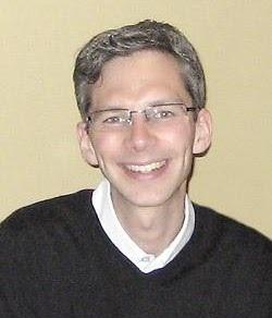 Ein lächelnder Mann mit Brille und grauen Haaren trägt einen schwarzen Pullover vor einer einfarbigen Wand.