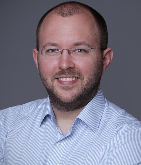 Portrait eines Mannes mit Brille und Bart, der lächelnd in einem hellblauen Hemd vor einem grauen Hintergrund steht.