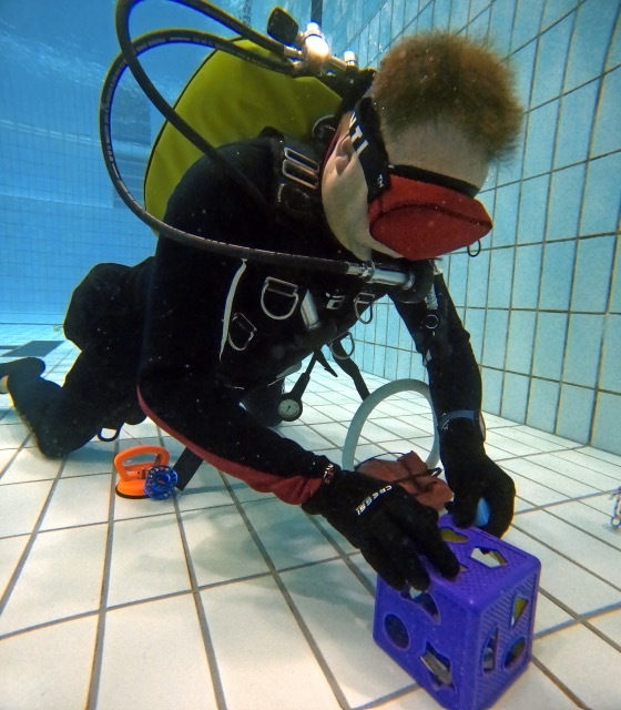 Taucher in schwarzer Ausr&uuml;stung untersucht einen bunten W&uuml;rfel auf dem Boden eines Schwimmbeckens.