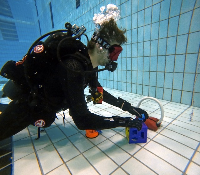 Taucher in Ausr&uuml;stung legt einen bunten Block auf den Fliesenboden eines Schwimmbeckens. Blasen steigen an die Oberfl&auml;che.