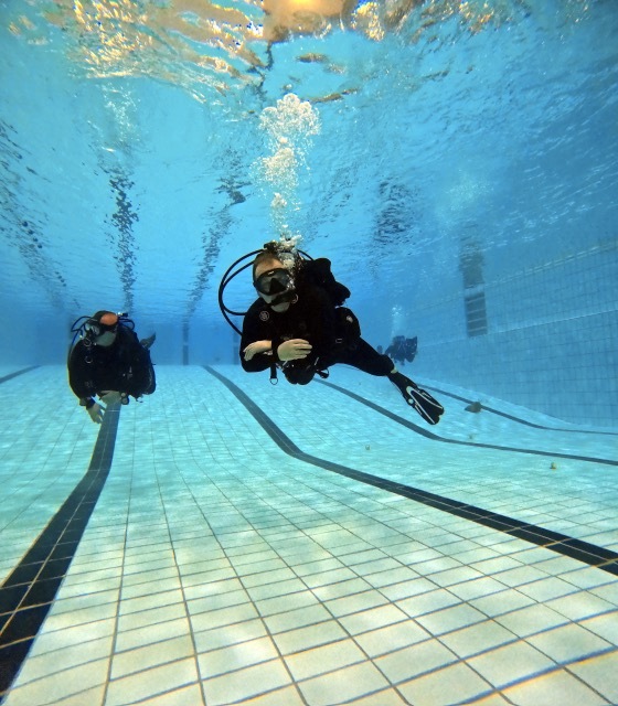 Unterwasseraufnahme zweier Taucher in Neoprenanz&uuml;gen, die &uuml;ber den gefliesten Grund eines Schwimmbads schwimmen.