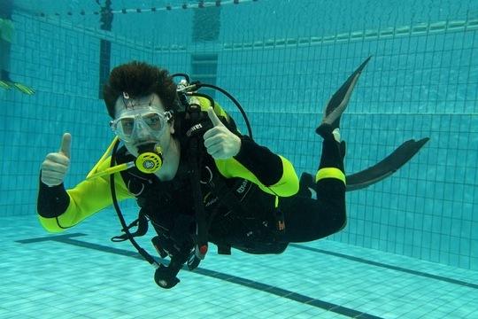 Taucher in einem Schwimmbecken zeigt Daumen hoch, tr&auml;gt Neoprenanzug und Atemger&auml;t, umgeben von blauen Fliesen.