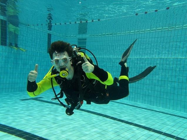 Taucher in einem Schwimmbecken zeigt Daumen hoch, trägt Neoprenanzug und Atemgerät, umgeben von blauen Fliesen.