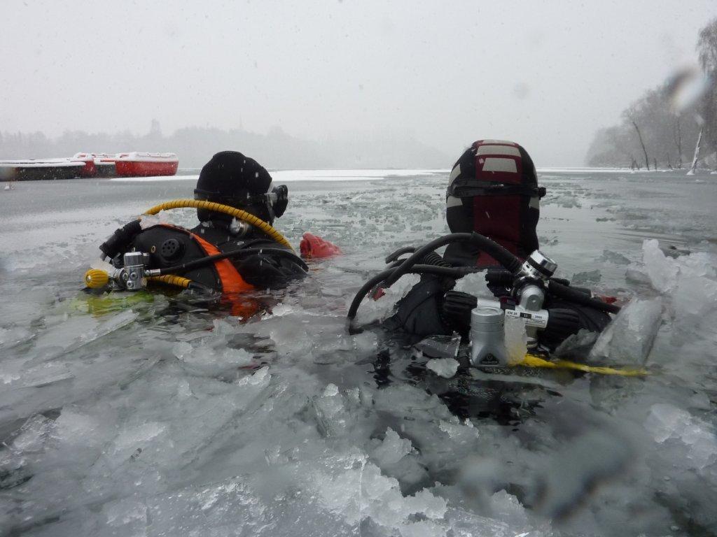 Zwei Taucher im Eiswasser, umgeben von Eisschollen, tragen Tauchausrüstung und schwimmen in einem gefrorenen Gewässer.