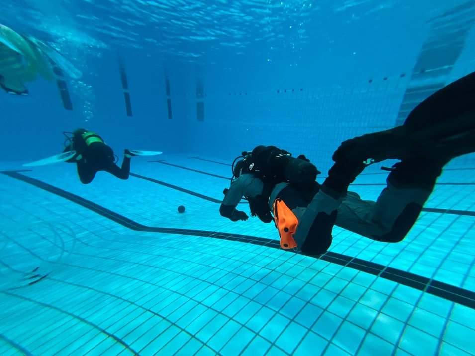 Zwei Taucher in Neoprenanz&uuml;gen schwimmen unter Wasser in einem Schwimmbad mit sichtbarem Fliesenboden.