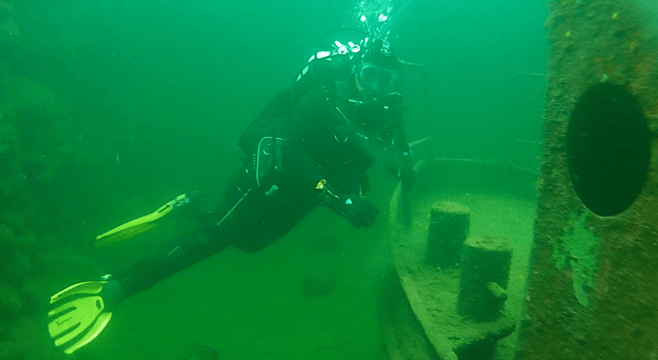 Taucher in schwarzer Ausrüstung erkundet ein versunkenes Schiffswrack in trübem Wasser mit grünem Farbton.