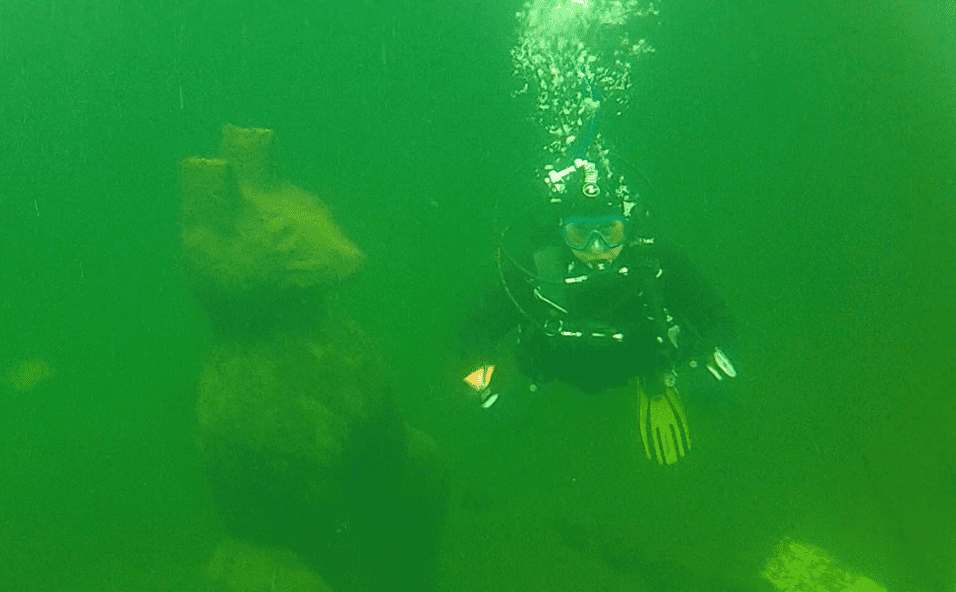 Taucher schwebt in trübem Wasser neben einer unter Wasser befindlichen Steinskulptur.