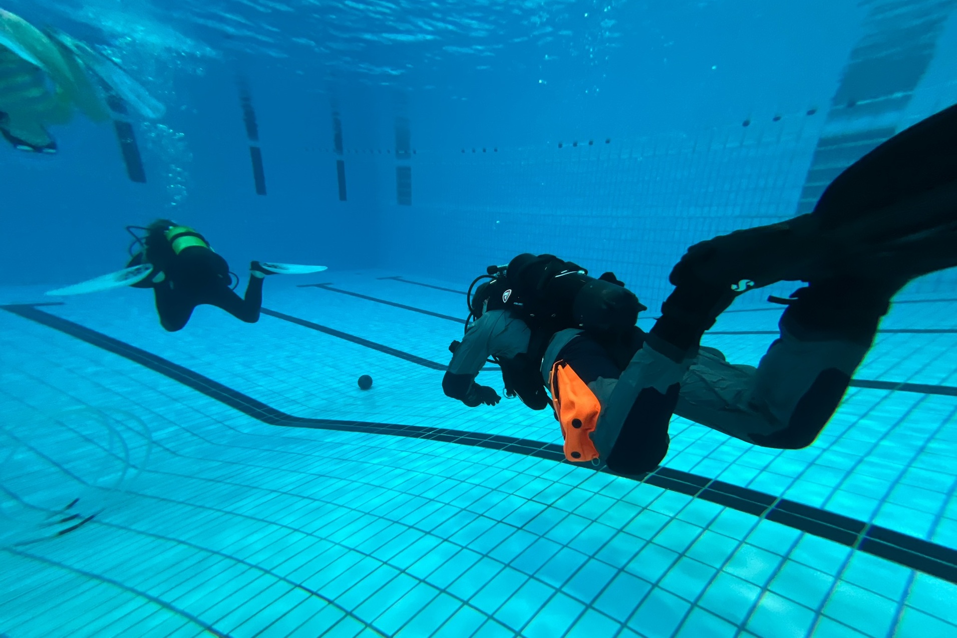 Zwei Taucher in Neoprenanz&uuml;gen schwimmen unter Wasser in einem Schwimmbad mit sichtbarem Fliesenboden.