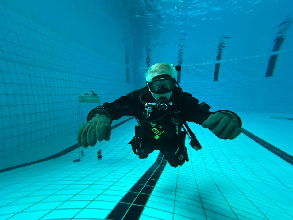 Taucher in schwarzem Neoprenanzug schwebt unter Wasser in einem Schwimmbecken mit blauen Fliesen.