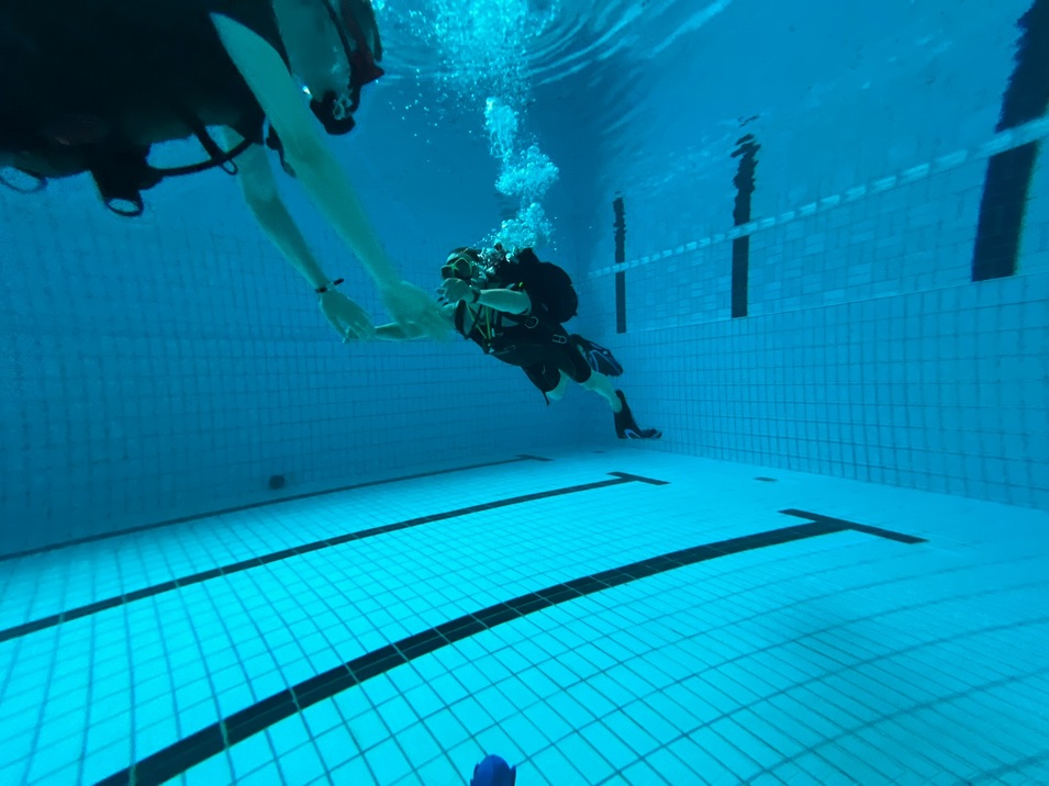 Taucher in einem Schwimmbad, unter Wasser, mit Ausr&uuml;stung und Luftblasen sowie Fliesenboden sichtbar.