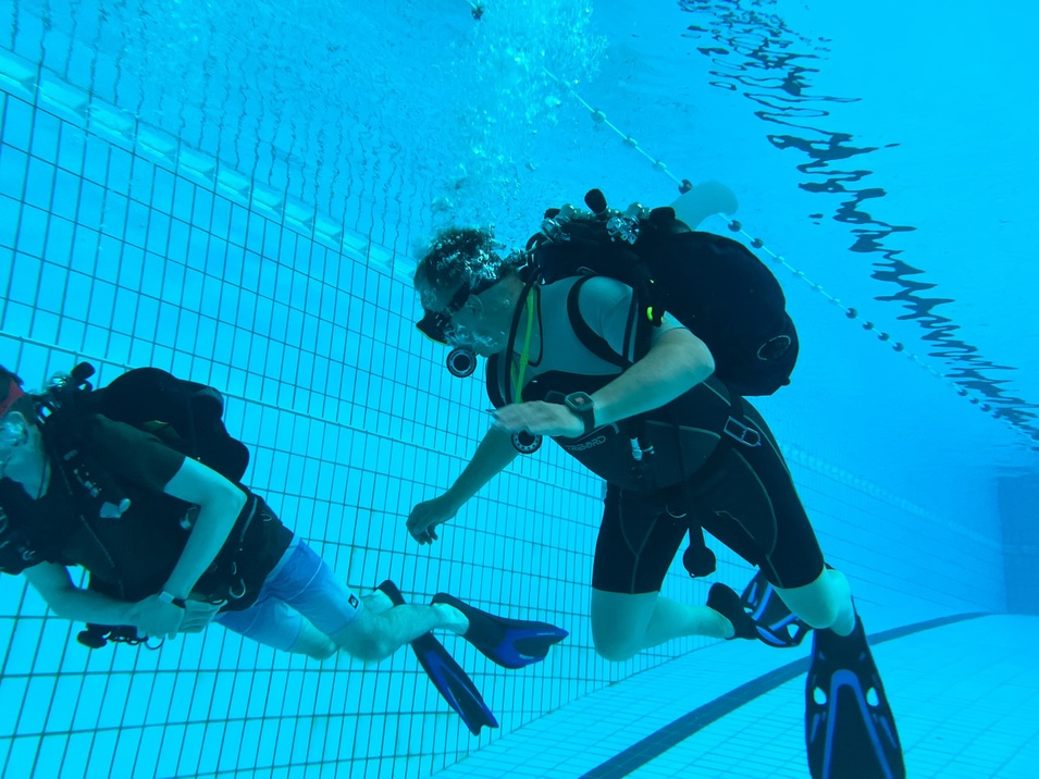 Zwei Taucher schwimmen unter Wasser in einem Schwimmbecken, beide mit Tauchanz&uuml;gen und Ausr&uuml;stung.