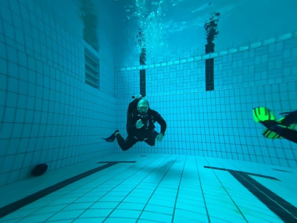 Taucher in Schwarz sitzt unter Wasser in einem Schwimmbecken mit blauen Fliesen und sprudelnden Blasen um ihn.