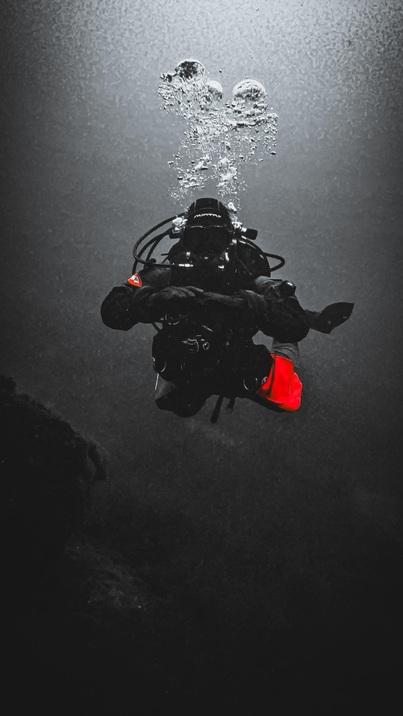 Taucher mit schwarz-gelber Ausrüstung schwebt unter Wasser, umgeben von Blasen und dunklem Hintergrund.