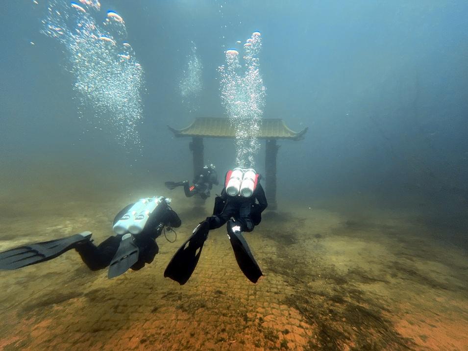 Zwei Taucher bewegen sich unter Wasser in der Nähe eines chinesischen Tempels im Hintergrund. Blasen steigen auf.