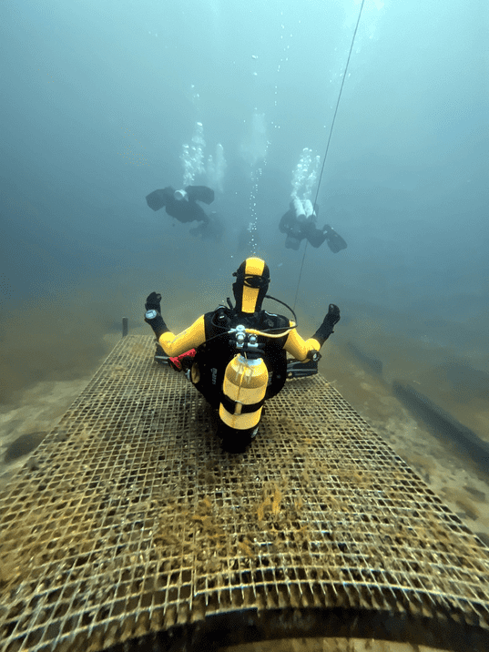 Taucher auf einem Gitterboden unter Wasser, während zwei weitere Taucher im Hintergrund aufsteigen.