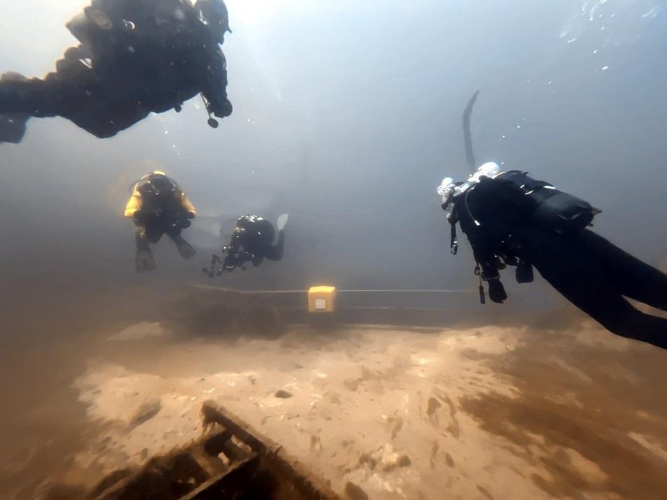 Fünf Taucher schwimmen unter Wasser über einem gesunkenen Wrack auf einem sandigen Meeresboden.