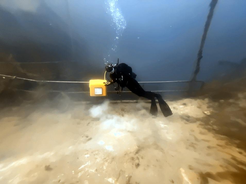 Taucher schwimmt unter Wasser in der Nähe eines gelben Geräts auf dem sandigen Grund. Blasen steigen um ihn herum auf.