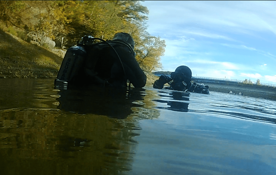 Zwei Taucher in schwarzer Ausrüstung arbeiten im flachen Wasser unter blauem Himmel.