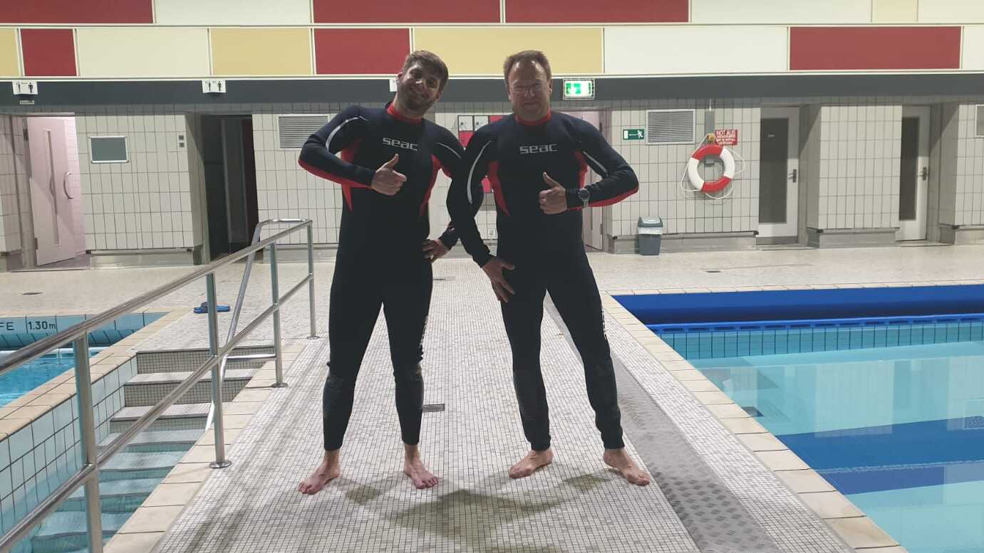 Zwei Schwimmer in Neoprenanzügen posieren am Beckenrand eines Hallenbades. Im Hintergrund ist ein Schwimmbecken sichtbar.