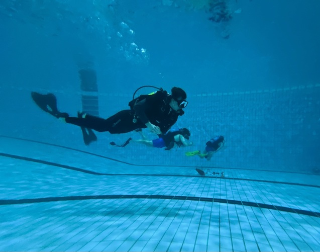 Taucher in schwarzer Ausr&uuml;stung schwimmt unter Wasser, begleitet von zwei weiteren Personen in Schwimmbekleidung.