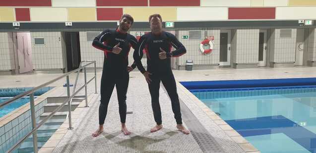 Zwei Schwimmer in Neoprenanzügen posieren am Beckenrand eines Hallenbades. Im Hintergrund ist ein Schwimmbecken sichtbar.