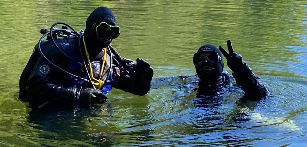 Zwei Taucher in Neoprenanzügen stehen im Wasser und zeigen ein Friedenszeichen, umgeben von ruhiger Wasseroberfläche.