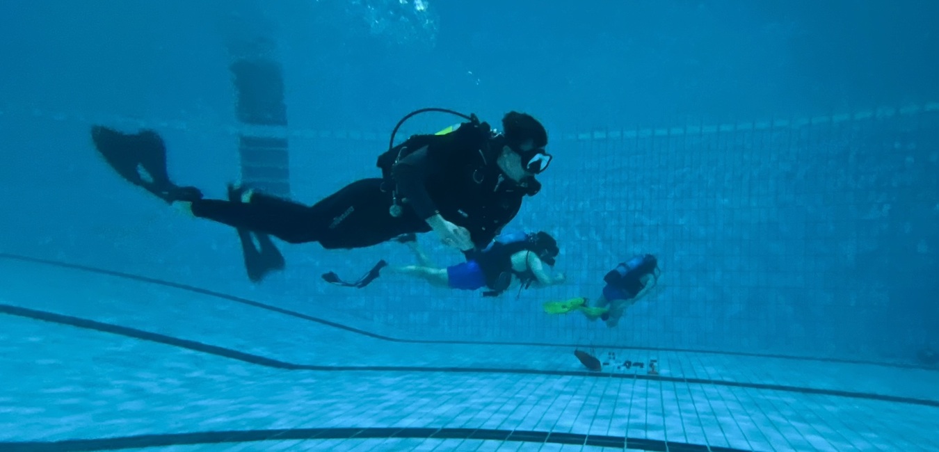 Taucher in schwarzer Ausr&uuml;stung schwimmt unter Wasser, begleitet von zwei weiteren Personen in Schwimmbekleidung.