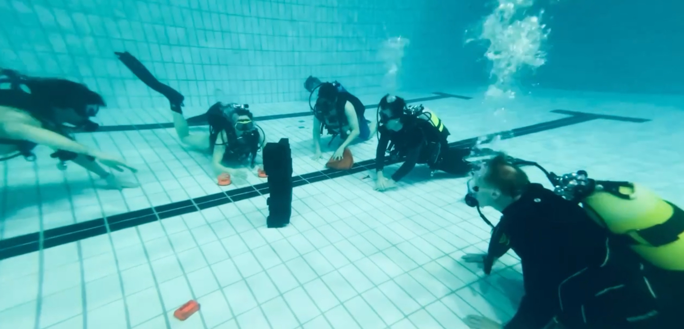 Sechs Taucher in Neoprenanz&uuml;gen &uuml;ben im Gew&auml;sser eines Hallenbades, umgeben von blauer Wasseroberfl&auml;che.