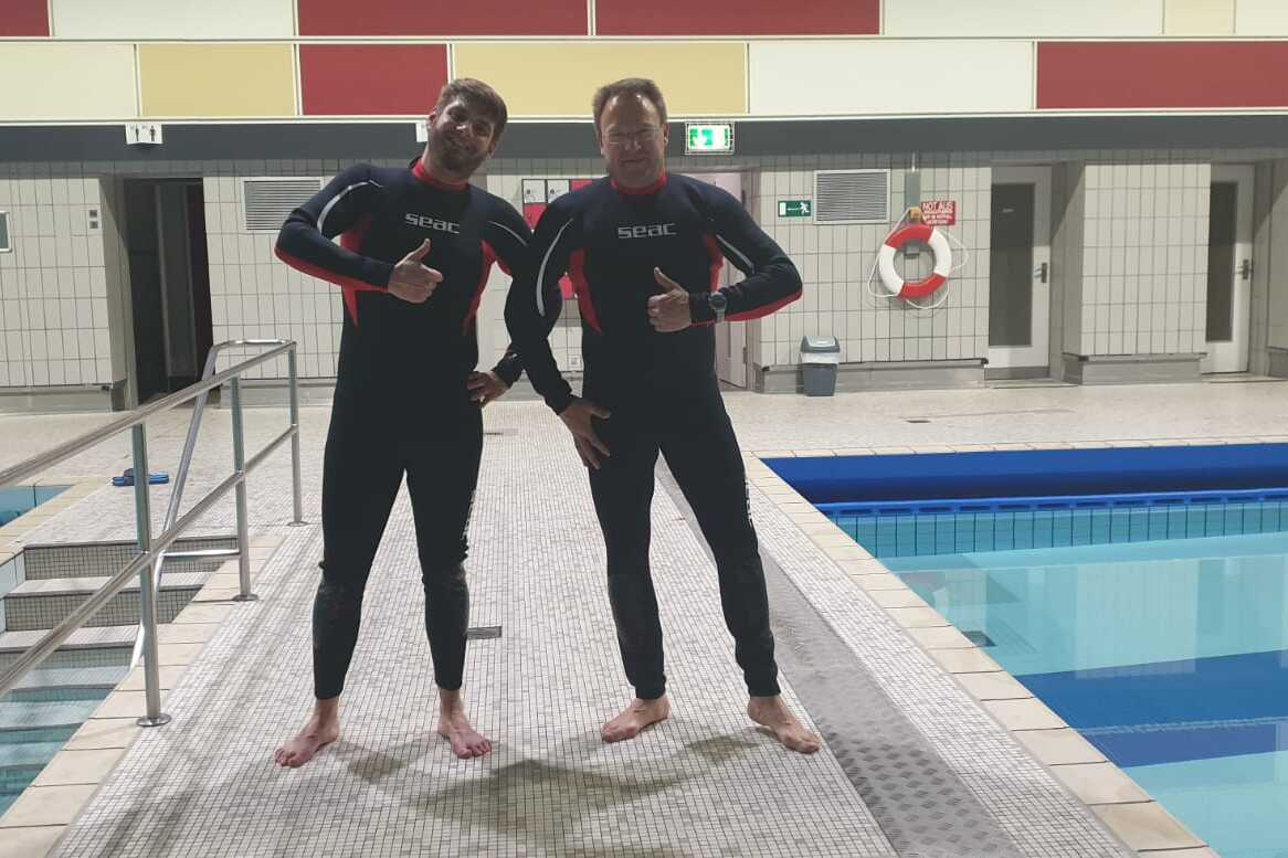 Zwei Schwimmer in Neoprenanz&uuml;gen posieren am Beckenrand eines Hallenbades. Im Hintergrund ist ein Schwimmbecken sichtbar.