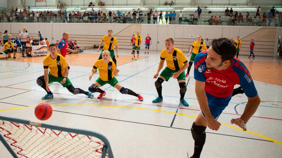 Spieler in blauer Trikots uniform wirft einen Ball auf ein Tor, w&auml;hrend vier Spieler in gelben Trikots bereitstehen.