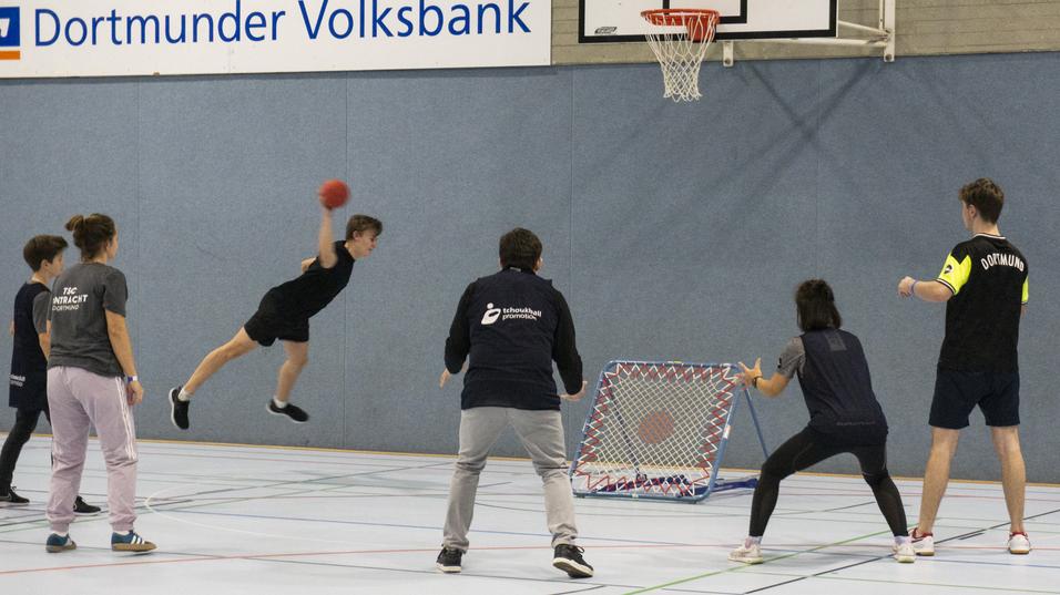Jugendlicher springt zum Torwurf in einer Sporthalle, umgeben von Spielern und einem Tor mit Netz.
