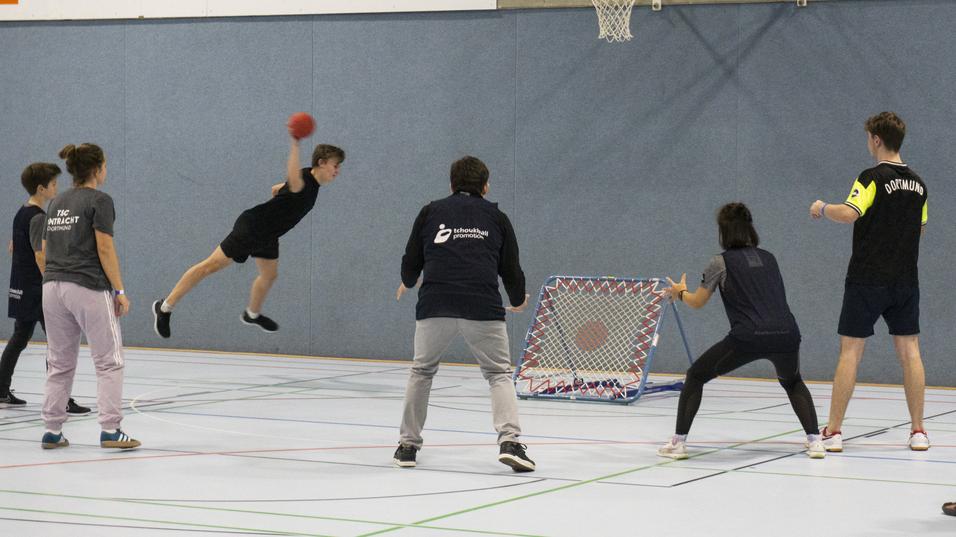 Jugendlicher springt zum Torwurf in einer Sporthalle, umgeben von Spielern und einem Tor mit Netz.