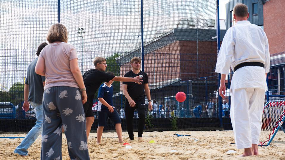 Gruppe von Menschen spielt ein Ballspiel im Sand, w&auml;hrend eine Person in Karate-Gewandung zuschaut.