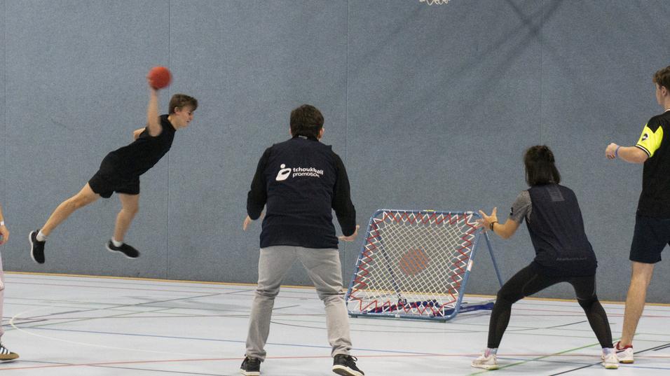 Jugendlicher springt zum Torwurf in einer Sporthalle, umgeben von Spielern und einem Tor mit Netz.