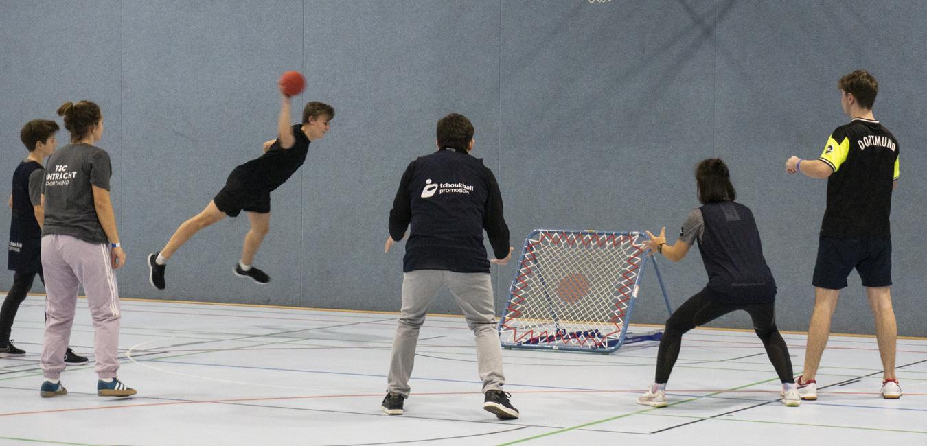 Jugendlicher springt zum Torwurf in einer Sporthalle, umgeben von Spielern und einem Tor mit Netz.