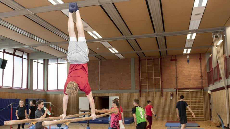 Ein Turner f&uuml;hrt einen Handstand an einem Barren in einer Sporthalle mit weiteren &Uuml;bungen im Hintergrund aus.