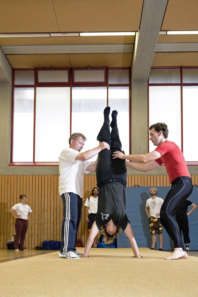 Drei Personen in sportlicher Kleidung üben Handstandtechniken in einer Turnhalle mit Sicht auf Fensterscheiben.