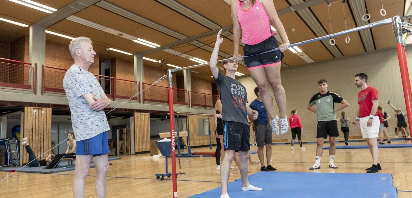 Sportliche Trainingseinheit in der Halle: Eine junge Athletin schwingt an einem Reck, während Trainer und Zuschauer zuschauen.
