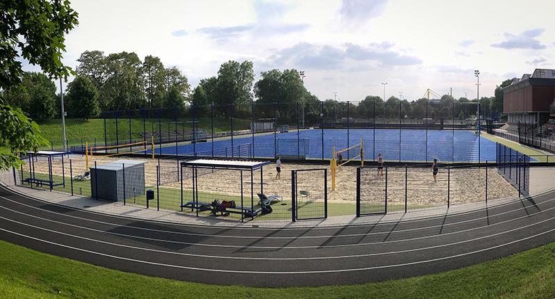 Sportanlage mit Sandvolleyballfeld und Tennisplätzen, umgeben von einem Zaun und einem Laufweg. Wolkiger Himmel im Hintergrund.