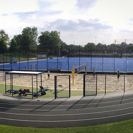Sportanlage mit Sandvolleyballfeld und Tennispl&auml;tzen, umgeben von einem Zaun und einem Laufweg. Wolkiger Himmel im Hintergrund.
