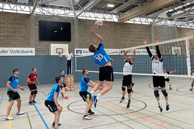 Volleyballspiel in einer Halle: Ein Spieler springt hoch, um einen Ball &uuml;ber das Netz zu schlagen, w&auml;hrend andere Spieler reagieren.