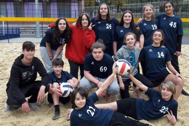 Gruppe von zw&ouml;lf Jugendlichen in Sportkleidung auf einem Sandvolleyballplatz, umgeben von Volleyballen und einem Netz.