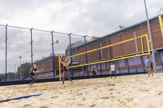 Strandvolleyballspiel mit mehreren Spielerinnen auf einem Sandplatz unter einem bew&ouml;lkten Himmel.