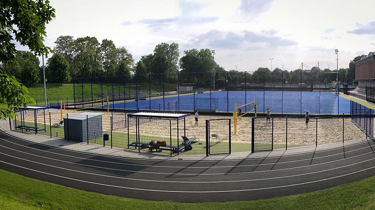 Sportanlage mit Sandvolleyballfeld und Tennispl&auml;tzen, umgeben von einem Zaun und einem Laufweg. Wolkiger Himmel im Hintergrund.