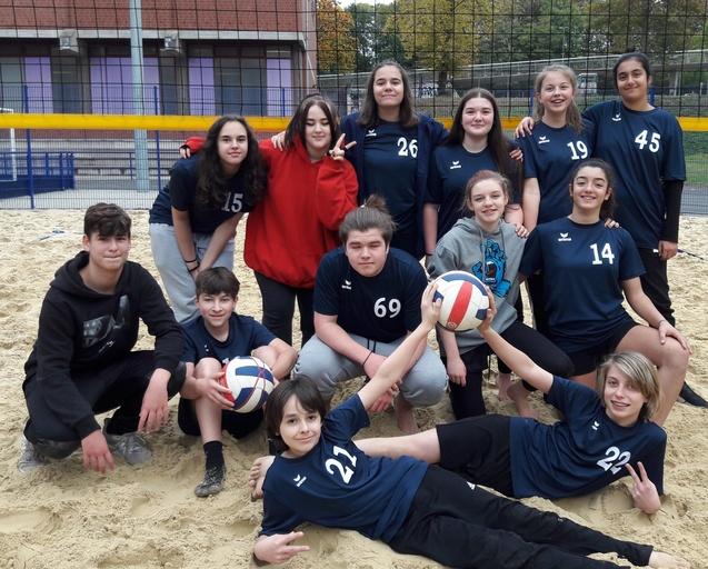 Gruppe von zwölf Jugendlichen in Sportkleidung auf einem Sandvolleyballplatz, umgeben von Volleyballen und einem Netz.