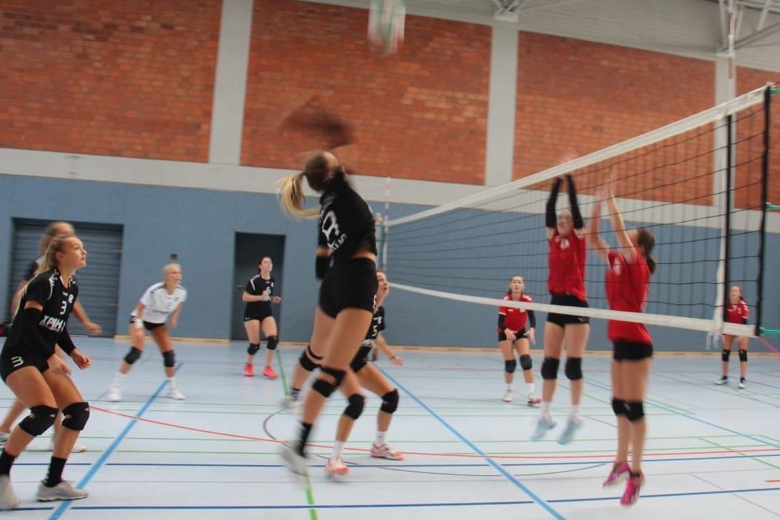 Volleyballspiel in einer Halle: Eine Spielerin springt zum Aufschlag, während sich Gegnerinnen zur Abwehr positionieren.